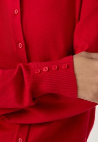 Cárdigan rojo hecho de un material suave, con un frente abotonado y puños acanalados con cinco botones a juego. Textura suave en todo.