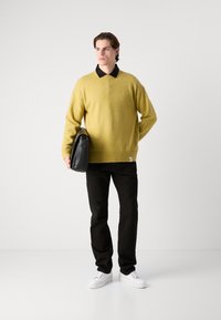 Gul strikket genser, svart skjorte med krage under, svarte bukser og hvite sneakers. Holder en svart skulderveske. Enkelt design.