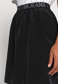 Falda negra plisada con cinturilla elástica que presenta el texto "CALVIN KLEIN JEANS" en blanco. Textura suave, diseño hasta la rodilla.