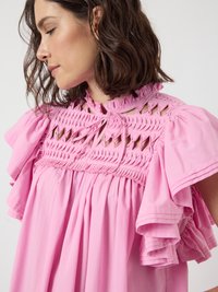 Sieviete ar plecu garumu brūniem matiem, ģērbta brīvā rozā kleitā ar sarežģītiem izgriezumiem un volāniem augšējā daļā un piedurkņās.