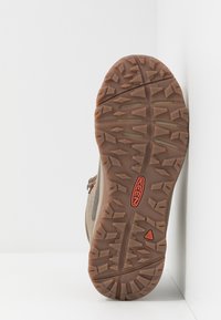 Suola di scarpa da trekking marrone chiaro con scanalature profonde. Presenta un fondo in gomma con presa testurizzata e un logo rosso. Dettagli di design minimi.