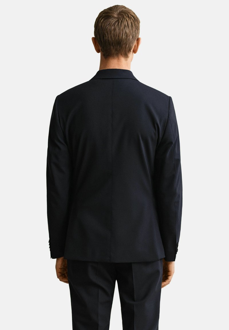 Homme aux cheveux châtains courts portant un costume noir ajusté, debout de dos sur un fond clair uni.