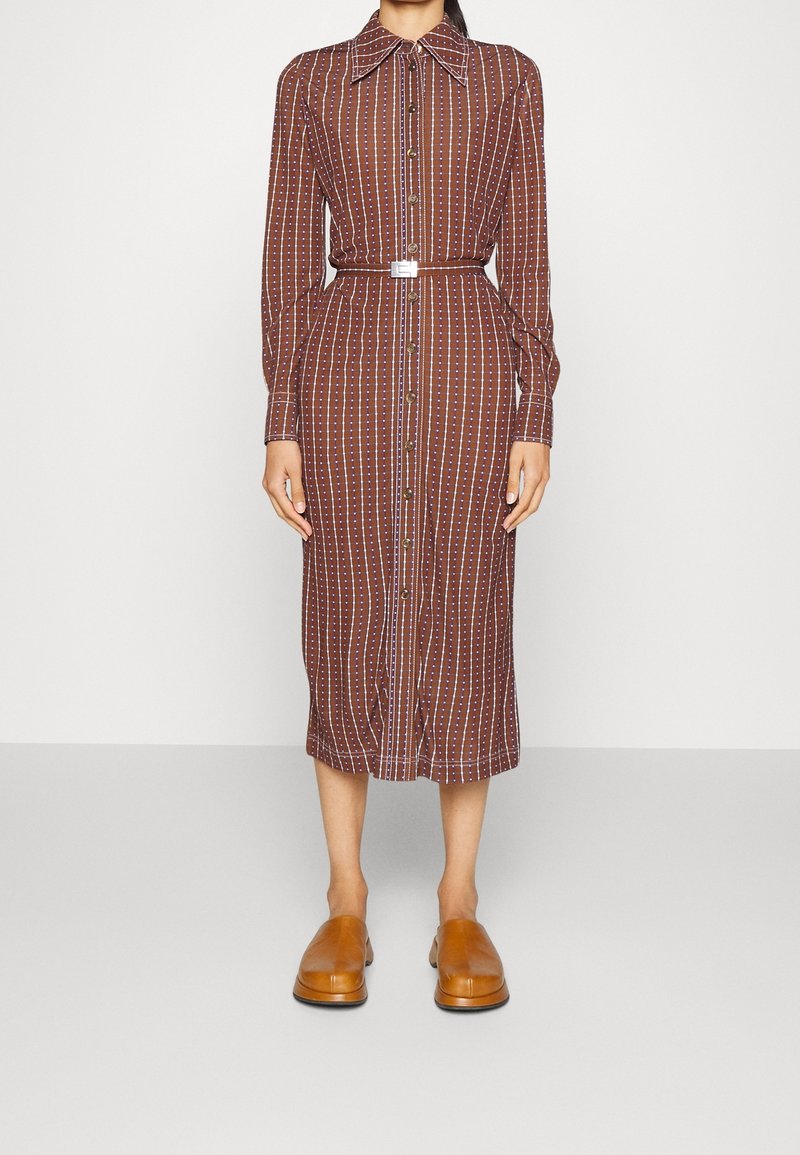 Femme portant une robe midi marron boutonnée avec des rayures blanches verticales, une ceinture assortie et des chaussures marron à enfiler, debout devant un fond blanc.