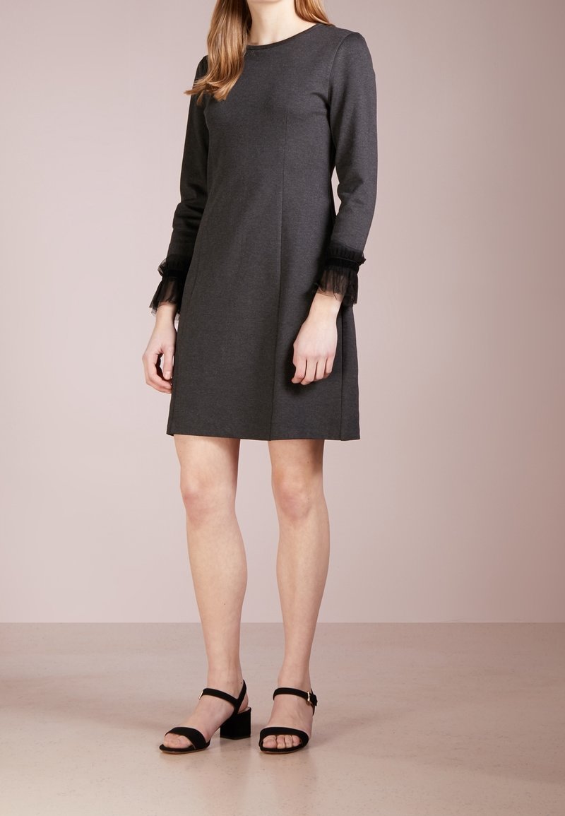 Grå klänning med långa ärmar, med svarta tyllaccenter vid ärmsluten. Rakt snitt och knä längd, kombinerad med svarta klacksandaler.