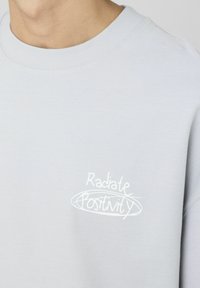 Sweatshirt gris clair à col rond et poignets côtelés. Présente un texte brodé blanc "Radiate Positivity" dans un design courbé.