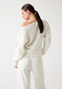 Pull en tricot blanc avec un design à épaule dénudée, couture détaillée le long de l'encolure, associé à un pantalon blanc taille haute avec des poches texturées.