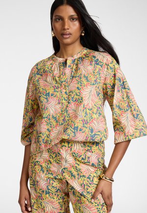 Vrouw met lang donker haar draagt bijpassende bloemen top en broek met grote roze en gele bladpatronen, geaccentueerd met gouden oorbellen en armband.