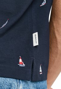Polo navy con motivo di barche a vela ricamate in rosso e bianco. Presenta uno spacco laterale e un'etichetta con il marchio "GANT" vicino all'orlo.