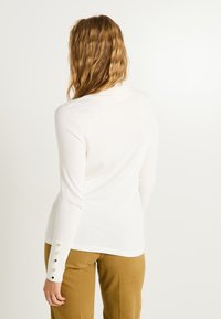 Top blanco de manga larga con escote redondo y detalles de botones en los puños, combinado con pantalones verde oliva. Textura suave y lisa.