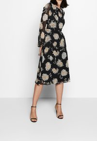 Svart midi-klänning med blommönster i beige och vitt. Genomskinliga långärmar, rund halsringning och volang i fållen. Bärs med svarta klacksandaler.
