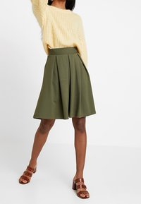 Olivgrön A-linjeformad kjol med veck, hög midja och knälång längd, kombinerad med bruna sandaletter med remmar och en krämfärgad stickad tröja.