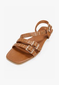 Sandaña de cuero marrón con tres correas ajustables que cuentan con tachuelas metálicas. Punta cuadrada y correa en el tobillo trasero con cierre de hebilla.