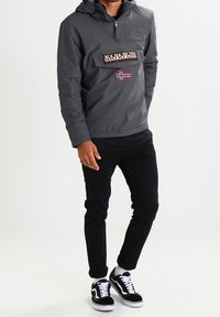 Mørkegrå letvægt anorakjakke med hætte, brystlomme og logo-patch; parret med sorte bukser og sort-hvide sneakers.