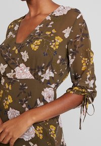 Robe portefeuille en tissu vert olive, ornée de motifs floraux roses et jaunes, avec des manches à nouer et une légère brillance.