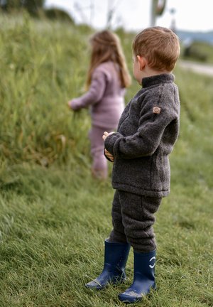 Vaikas vilki tamsiai pilką fliso aprangą ir mėlynas guminis batus, stovi ant žolės, netoliese yra mergaitė, apsirengusi šviesiai violetine apranga.