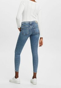 Vaqueros ajustados de mezclilla en color azul, con cintura alta, bolsillos traseros y un corte elegante. Combinados con zapatillas blancas y una blusa blanca texturizada.