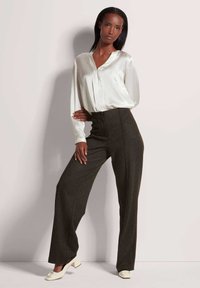 Weiße Seidenbluse mit langen Ärmeln, kombiniert mit hochgeschnittenen, dunkelbraunen weiten Hosen und weißen Schuhen mit niedrigem Absatz.