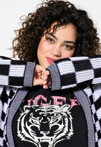 Gilet à carreaux noir et blanc avec tricot texturé, superposé sur une chemise noire avec un graphique de tigre blanc et des lettres roses.