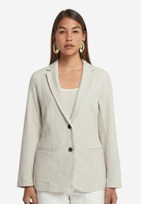 Ljusbeige blazer med ett texturerat mönster, enkelknäppt design, två framfickor och mörka knappar. Bärs över en vit topp.