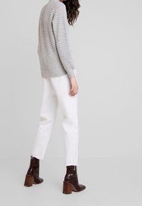 Pull gris tricoté avec un motif en zigzag, associé à un pantalon blanc à jambes droites et des bottes marron à talons, mettant en valeur un mélange de textures et de tonalités.