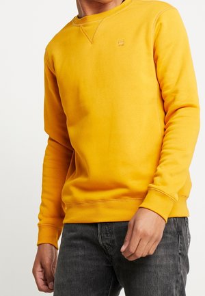 Homme portant un sweat-shirt jaune ajusté avec poignets et ourlet côtelés, associé à un jean noir délavé sur fond blanc.
