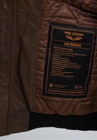 Giubbotto di volo in pelle marrone con texture trapuntata, polsini a costine, zip in metallo e un'etichetta che dettaglia le specifiche del prodotto.