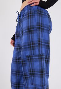 Blauwe geruite broek met zwarte accenten, voorzien van een elastische tailleband en zijzak. Materiaal lijkt zacht en textuur te hebben. Recht model.