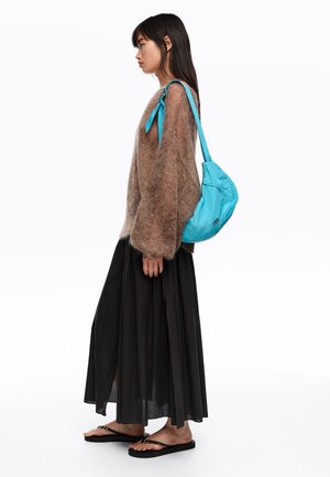 Mujer con suéter marrón de gran tamaño, falda larga negra, chanclas negras y llevando un bolso de hombro azul brillante.