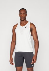 Weißes ärmelloses Sport-Tanktop mit Netzdesign und schwarzem Nike-Logo, kombiniert mit schwarzen enganliegenden Shorts mit weißem Logo.