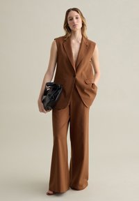 Bruine mouwloze blazer met één knoop, gecombineerd met wijde broek in dezelfde stof, vastgehouden met een zwarte tas. Gladde textuur.