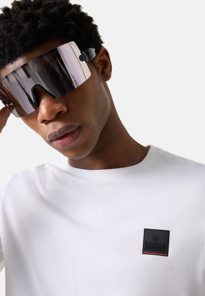 Jeune homme ajustant de grandes lunettes de soleil à visière réfléchissante, portant un t-shirt blanc avec un patch logo Fire+Ice noir et rouge.