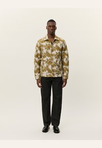 Long-sleeve, collar shirt in olive-green with white abstract patterns. Worn with black straight-leg trousers and black shoes.