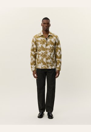 Long-sleeve, collar shirt in olive-green with white abstract patterns. Worn with black straight-leg trousers and black shoes.