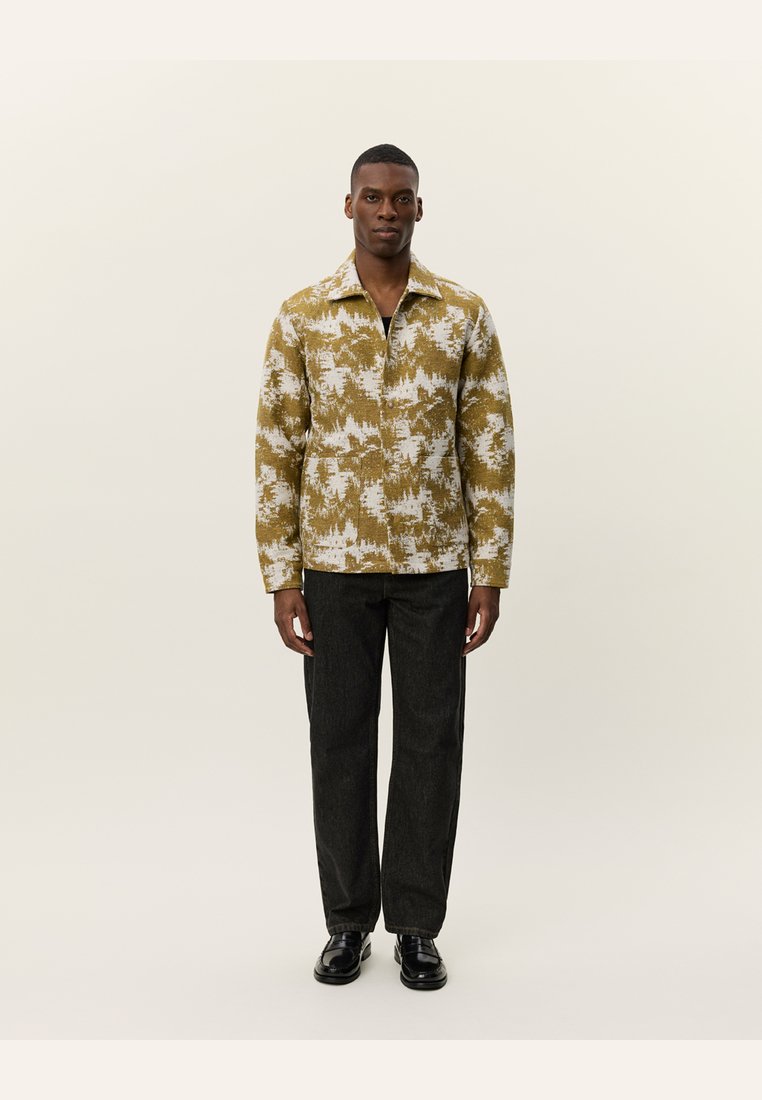 Long-sleeve, collar shirt in olive-green with white abstract patterns. Worn with black straight-leg trousers and black shoes.