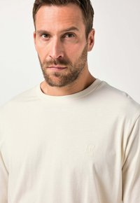 Cremefarvet langærmet t-shirt lavet af blødt stof, med et diskret præget logo på brystet og en rund halsudskæring.
