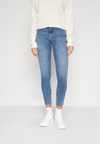 Jeans de denim azul ajustados con una cintura de altura media, que cuentan con un clásico diseño de cinco bolsillos, un sutil desgaste y un dobladillo a la altura del tobillo, combinados con zapatillas blancas.