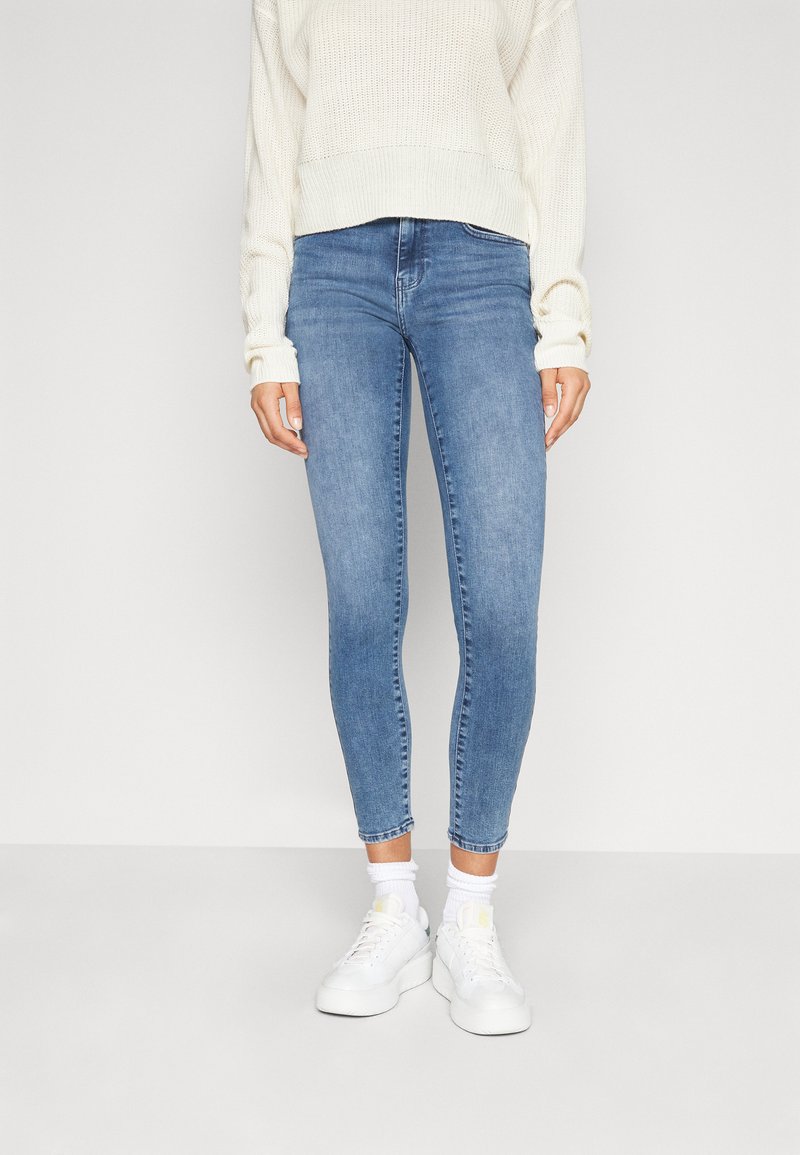 Jeans de denim azul ajustados con una cintura de altura media, que cuentan con un clásico diseño de cinco bolsillos, un sutil desgaste y un dobladillo a la altura del tobillo, combinados con zapatillas blancas.