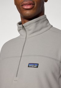 Sudadera de forro polar gris con cuello de cremallera al cuarto, que cuenta con un parche de logo negro de Patagonia en el pecho. Textura suave y diseño minimalista.
