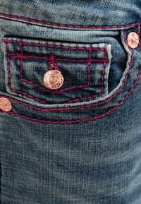 Dżinsowa kieszeń z kontrastującym bordowym przeszyciem, miedzianymi metalowymi elementami i małym haftowanym logo. Tło stanowi wyblakła niebieska faktura.