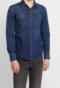 Mörkblå denimskjorta med långa ärmar, spetsig krage, två bröstfickor och knäppning fram. Sliten textur och rundad nederkant.