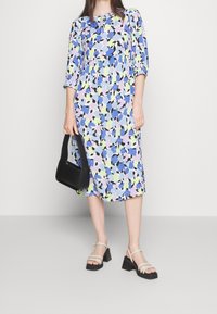 Bloemjurk in blauw, lavendel, groen en zwart met 3/4 mouwen en een losse pasvorm, gedragen met een zwarte schoudertas en blokhak sandalen.