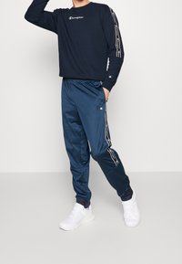 Marineblauw langshirt met wit logo, gecombineerd met marineblauwe trainingsbroek met zijstrepen en elastische tailleband. Witte sneakers maken de look af.