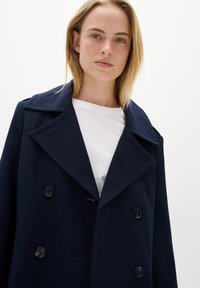 Palton bleumarin, cu două rânduri de nasturi, fabricat dintr-un material gros, cu rever mari și nasturi negri, purtat peste un tricou alb.