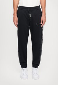Svarte joggebukser laget av myk stoff, med snøring i livet, sidestriper og brodert logo på forsiden. Kombinert med hvite sneakers.