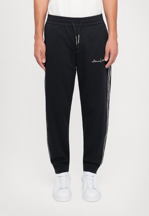 Zwarte joggingsbroek van zacht stof, met een trekkoord in de taille, zijkanten met strepen en een geborduurd logo op de voorkant. Gecombineerd met witte sneakers.