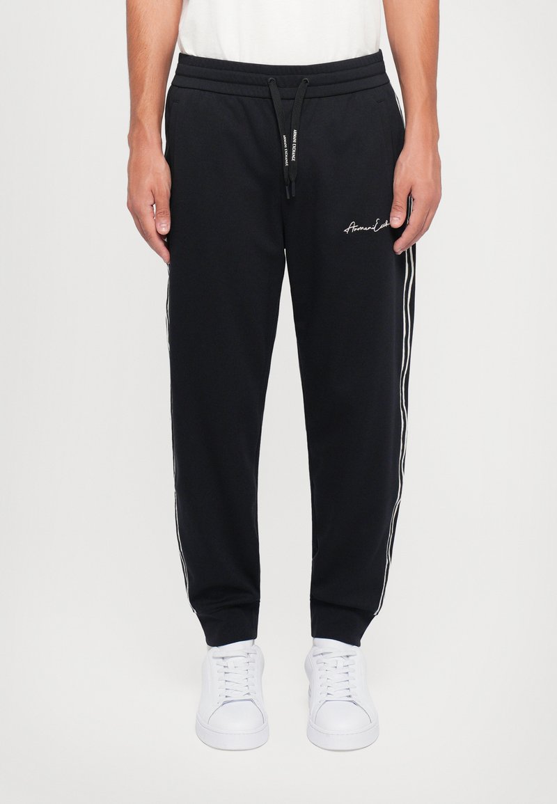 Svarte joggebukser laget av myk stoff, med snøring i livet, sidestriper og brodert logo på forsiden. Kombinert med hvite sneakers.