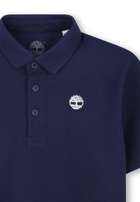 Polo bleu marine avec un col classique, trois boutons et un logo blanc représentant un arbre. Tissu texturé avec des manches courtes.