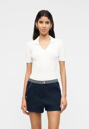 Vrouw met lang donker haar, gekleed in een wit geribd topje met korte mouwen en marineblauwe shorts met een gestreepte tailleband, die recht vooruit kijkt met een neutrale uitdrukking.