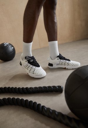 Ben iført hvide træningssko og sokker står på et fitnessgulv nær et sammenrullet kampreb og to sorte medicinbolde.