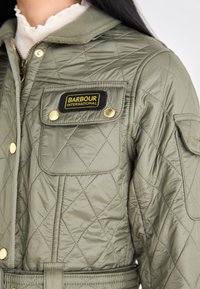 Olijfgroene gewatteerde jas met gouden hardware, voorzien van een zwart en geel Barbour International-embleem en zijkристи.